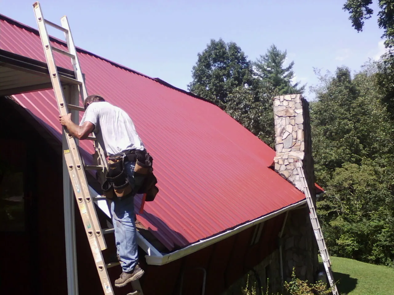 Roofer on a ladder beside a red metal roof and stone chimney — Soffit & Fascia Repair by Roofing Brothers in Freeman
